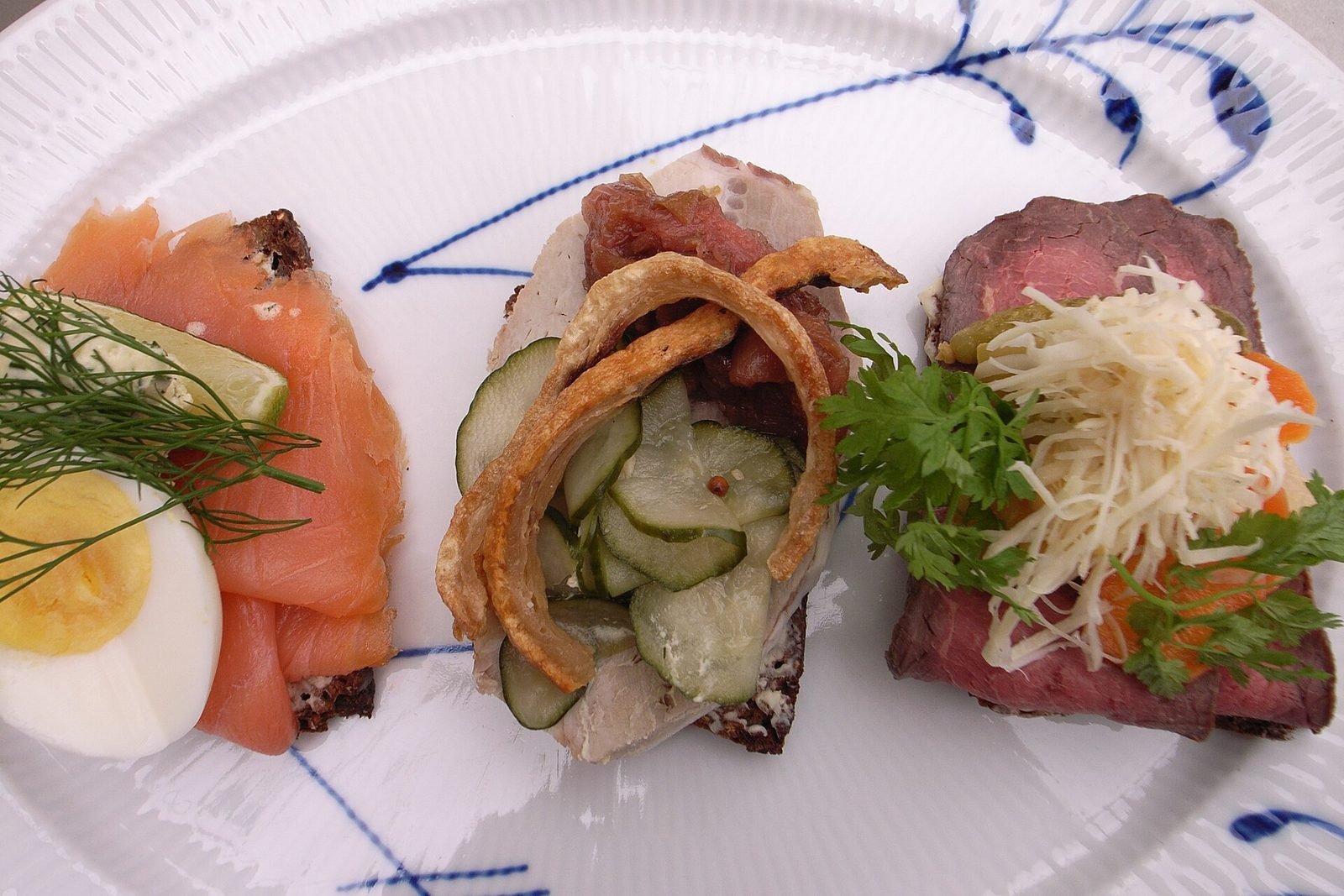 Drie belegde smørrebrød met zalm en ei, haring en rosbief op een blauw gedecoreerd bord